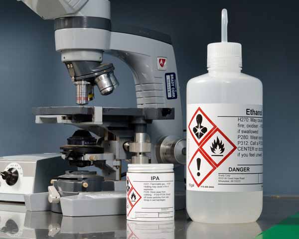 Un microscopio ubicado junto a botellas de alcohol isopropílico (IPA) y etanol, cada una con etiquetas del Sistema Globalmente Armonizado (GHS) para seguridad e identificación química.