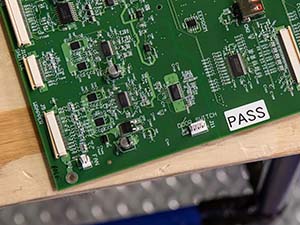 Placa de circuito electrónico con etiqueta de inspección 'PASS' colocada sobre una mesa de madera.