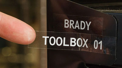Una etiqueta transparente con escritura blanca que dice Brady toolbox 01, aplicada a una pieza de metal.