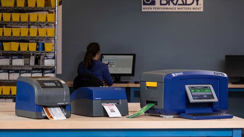 Tres diferente impresoras de etiquetas Brady mostrando etiqyetas de seguridad en una oficina.